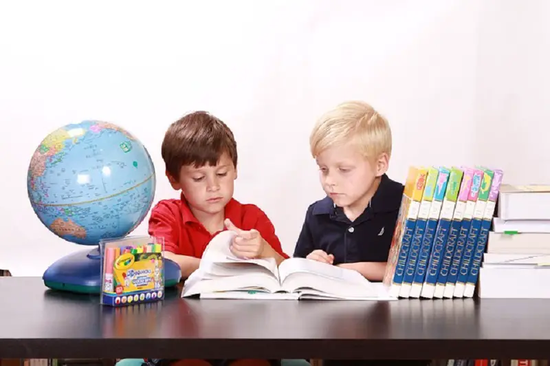 Two boys sitting at a table and reading a book. There are other books and a globe on the table.