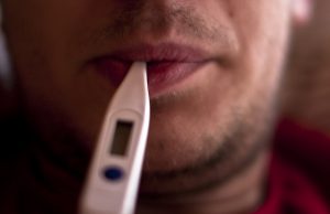 A lightly bearded man holding a thermometer in his mouth